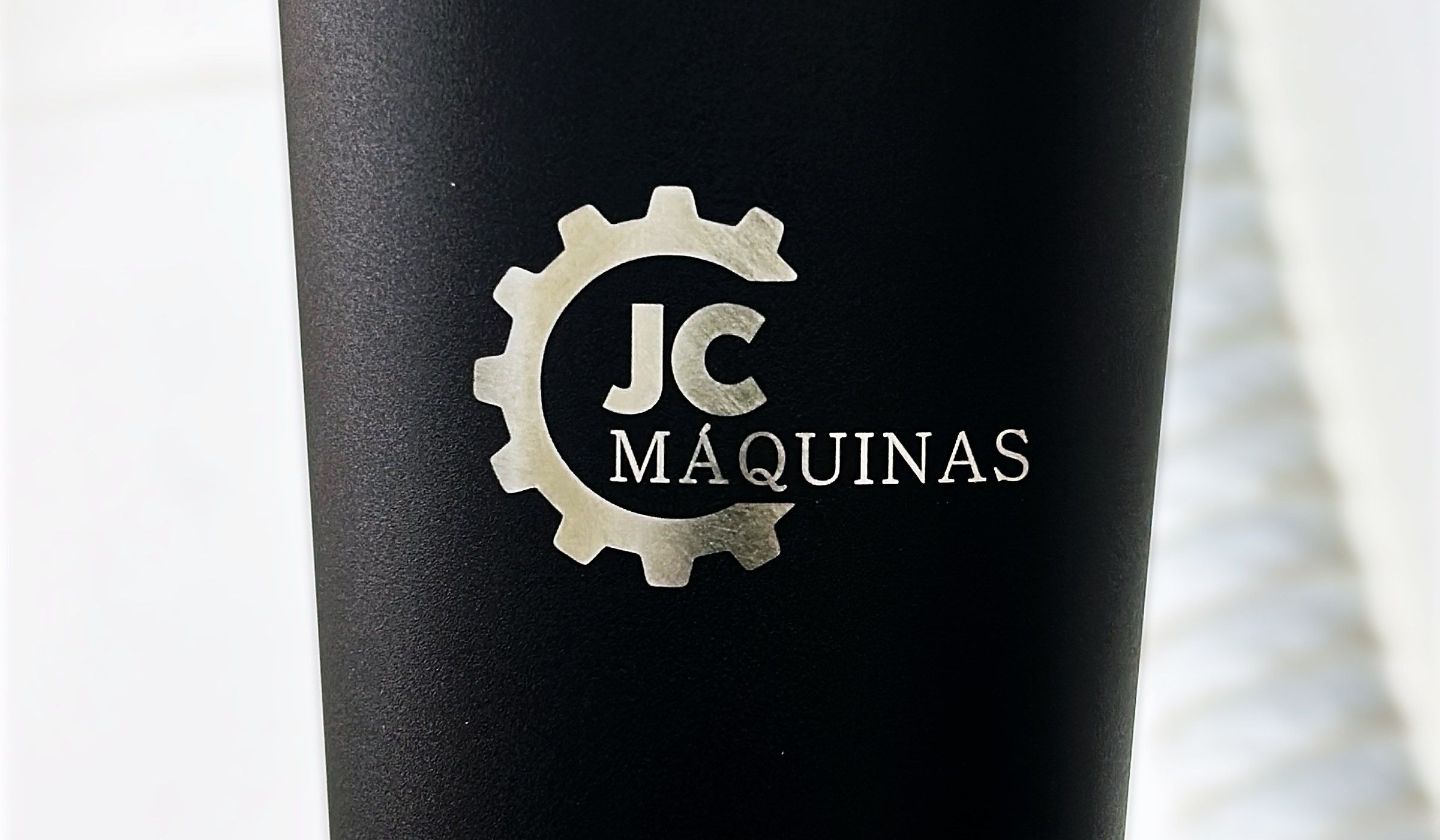 Copo térmico preto com o logo JC Máquinas gravado a laser, sobre a mesa da máquina