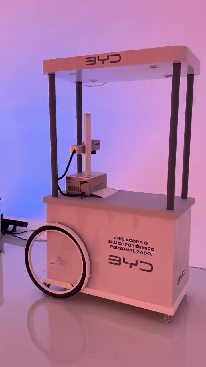 Estação de gravação a laser da Laser Design montada no estande da BYD, sob um toldo com a marca; a câmera se afasta e revela a estrutura de luz e o carro em exposição